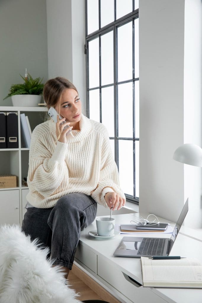 Female self employed professional working on a bench of her new lease to own home looking out the window while on the phone with her laptop and a hot beverage.