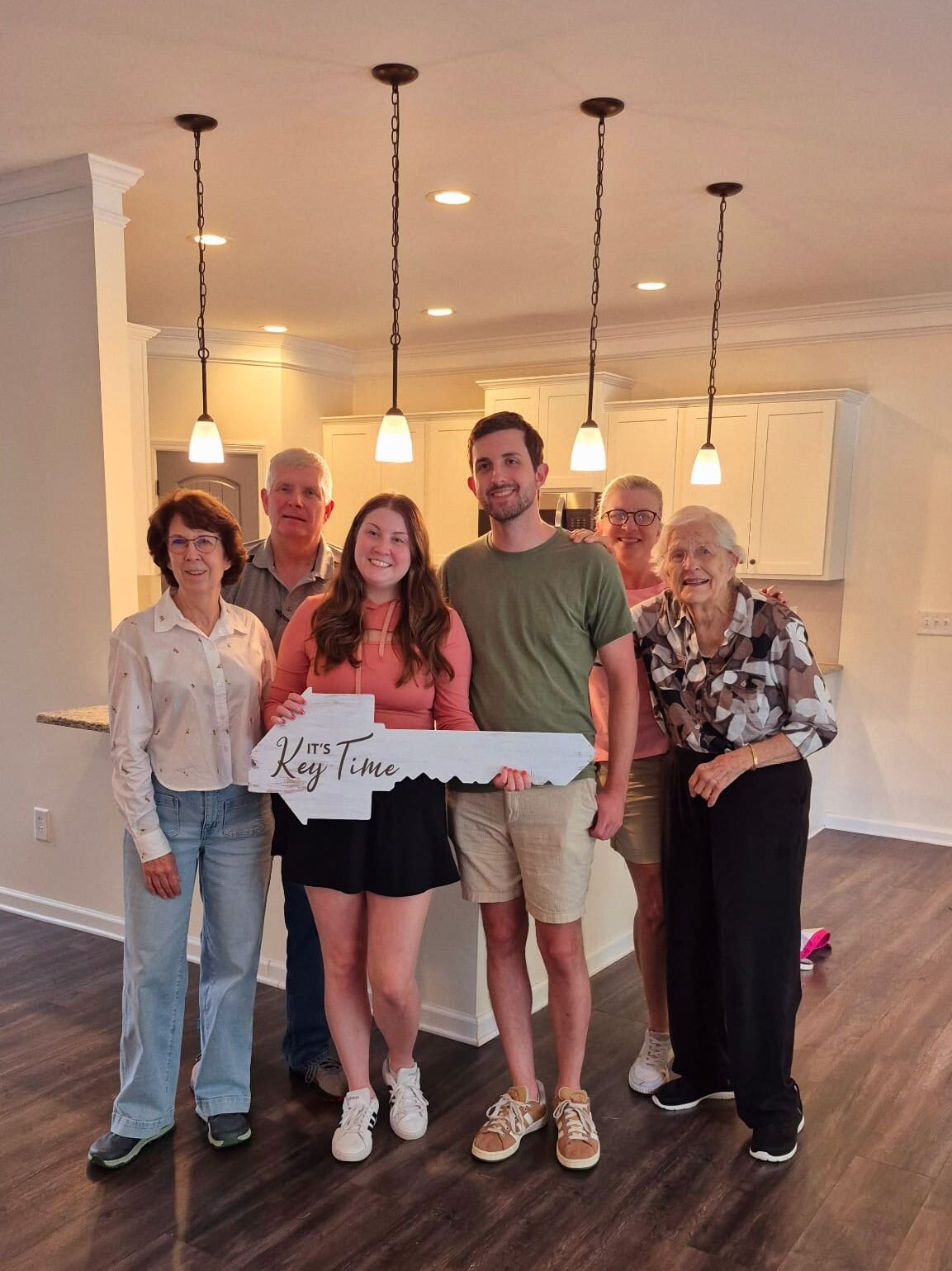 A group of six people stand smiling in a well-lit, new kitchen. The couple in the center holds a large key sign that reads "It's Key Time," symbolizing a joyous homecoming.