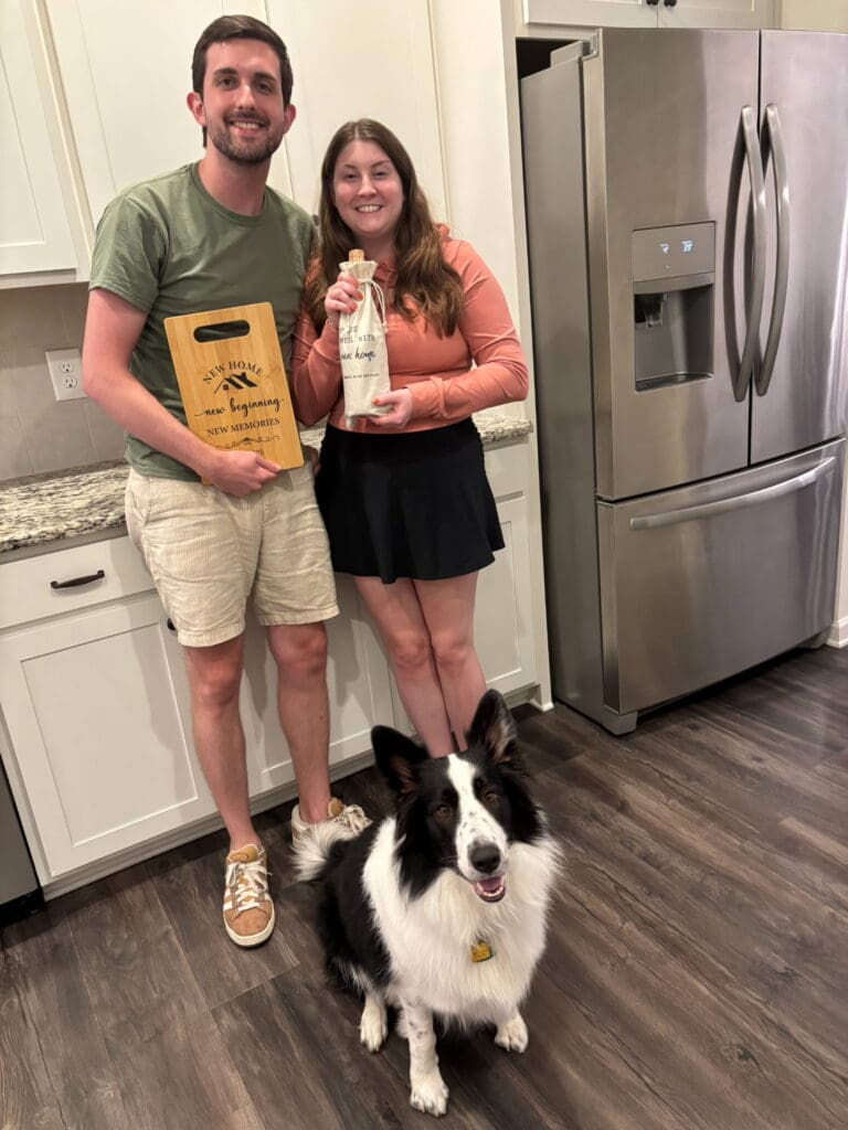 A smiling couple stands in a kitchen, holding a cutting board and a bottle labeled “new home.” A black and white dog sits in front of them.