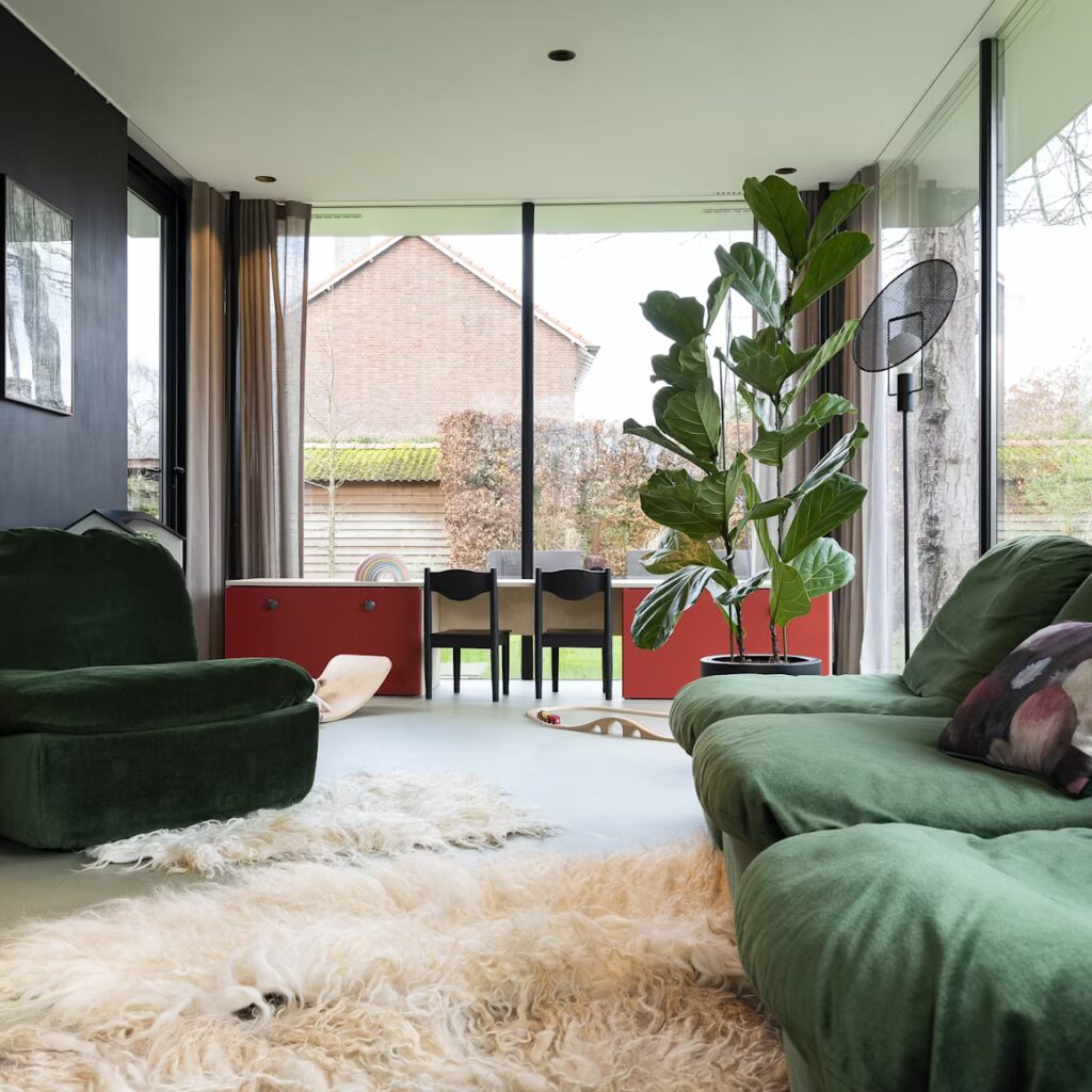 Modern living room with large windows, featuring green velvet couches and a plush white rug. A tall plant and floral vase add vibrancy. Cozy and inviting.