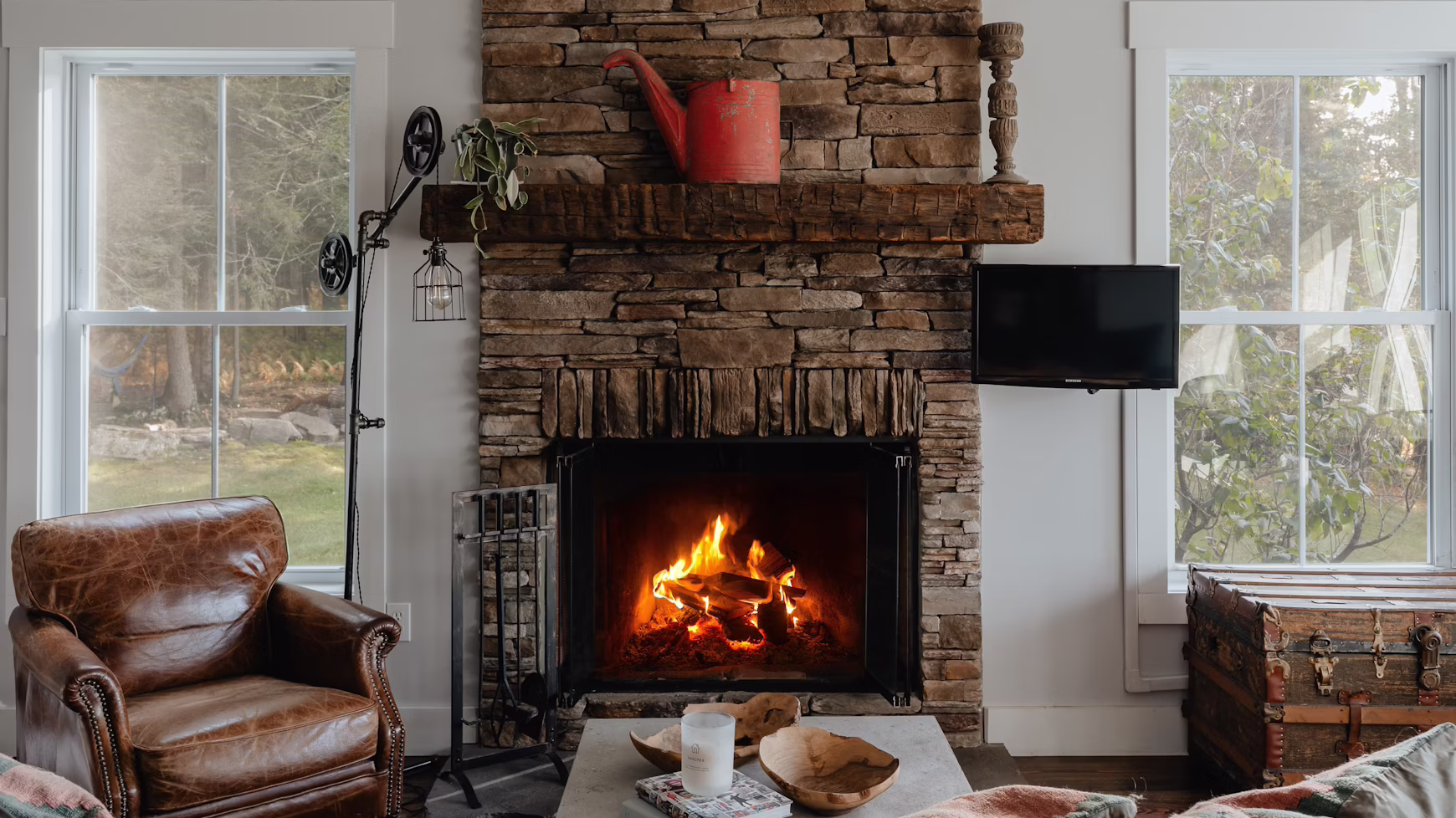 Cozy living room with a stone fireplace, lit fire, brown leather armchair, rustic decor, large windows, and a vintage trunk, evoking warmth.