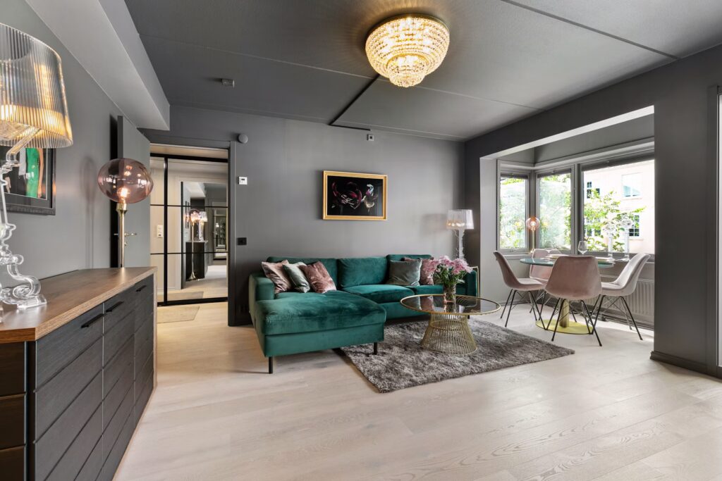 Chic living room with a green velvet sectional, gray walls, chandelier, glass coffee table, and dining area with pink chairs, evoking elegance and comfort.