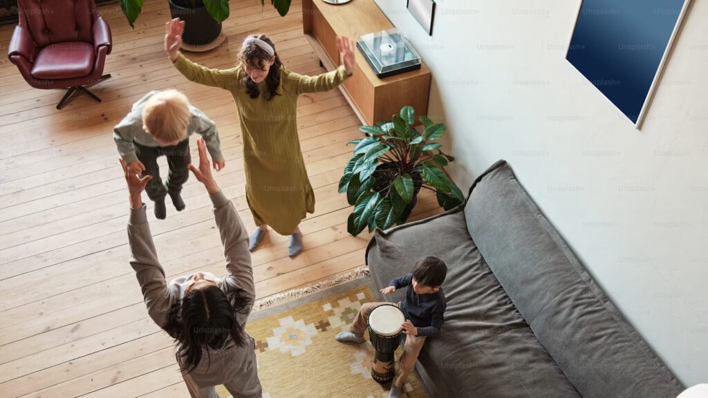 A joyful family scene in a living room with wooden flooring. An adult lifts a child into the air, another child plays a drum, and a woman watches excitedly.